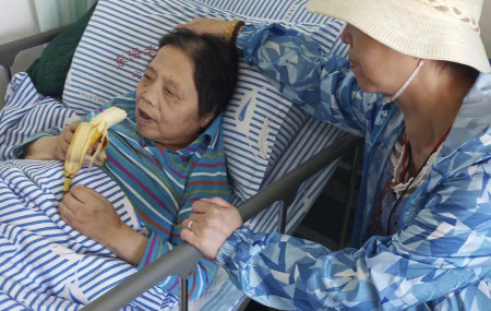 In this photo released by the family of Shen Peiming, Shen Peiming, 71, eats a banana as a family member attends to her at her bed side at the Shanghai Donghai Elderly Care hospital on Sept. 24, 2019.