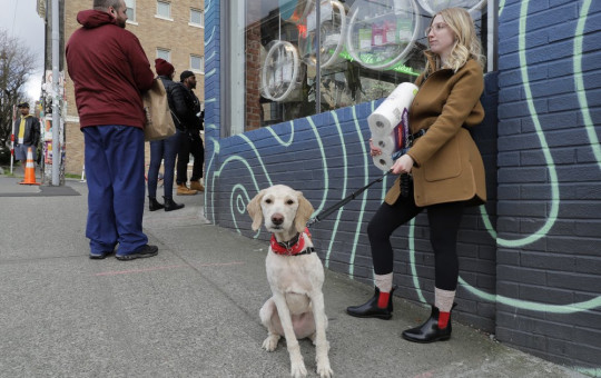 In this photo taken March 24, 2020, Mia Grace, right, holds a package of toilet paper as she and her dog Breezy observe social distancing chalk marks on the sidewalk while waiting to get in to The Reef Capitol Hill, a marijuana store in Seattle.