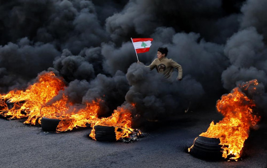 FILE- An anti-government demonstrator holds a national flag and runs across tires that were set on fire to block a main highway during a protest in the town of Jal el-Dib, north of Beirut, Lebanon, Tuesday, Jan. 14, 2020. AP/RSS Photo