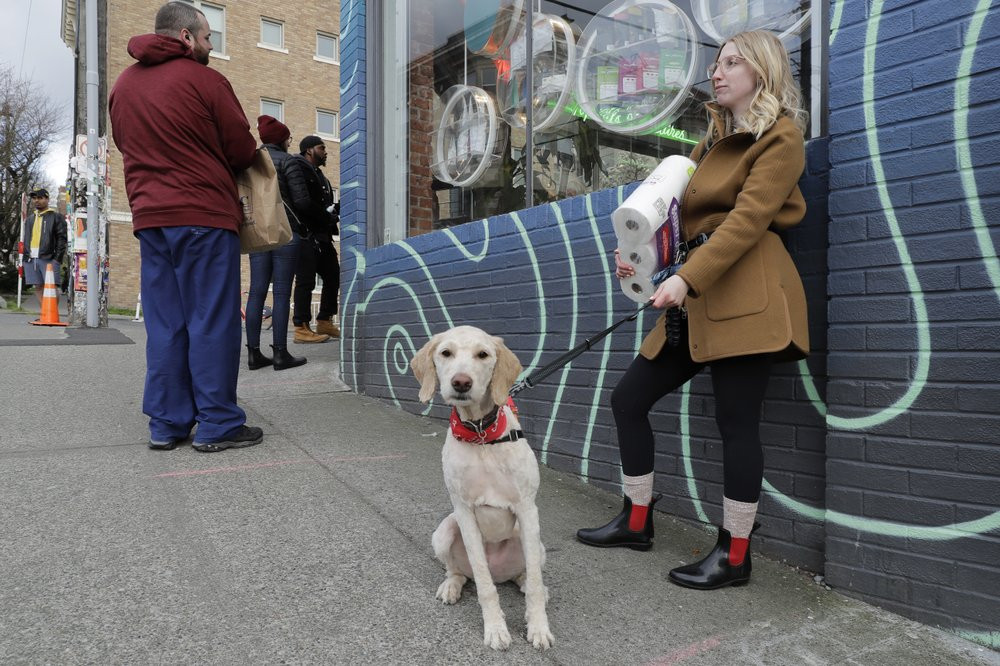 In this photo taken March 24, 2020, Mia Grace, right, holds a package of toilet paper as she and her dog Breezy observe social distancing chalk marks on the sidewalk while waiting to get in t