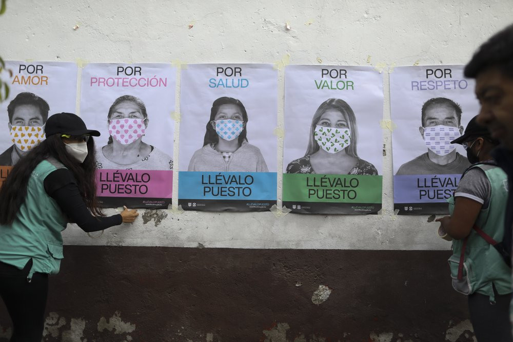 A man not wearing a face mask walks by as city health department workers affix a series of posters advocating mask use to a wall, in San Mateo Xalpa in the Xochimilco district of Mexico City,
