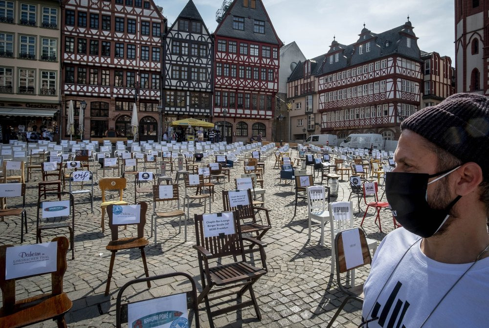 In this April 24, 2020 file photo, a man with a face mask watches empty chairs with names of bars and restaurants on the Roemerberg square in Frankfurt, Germany.