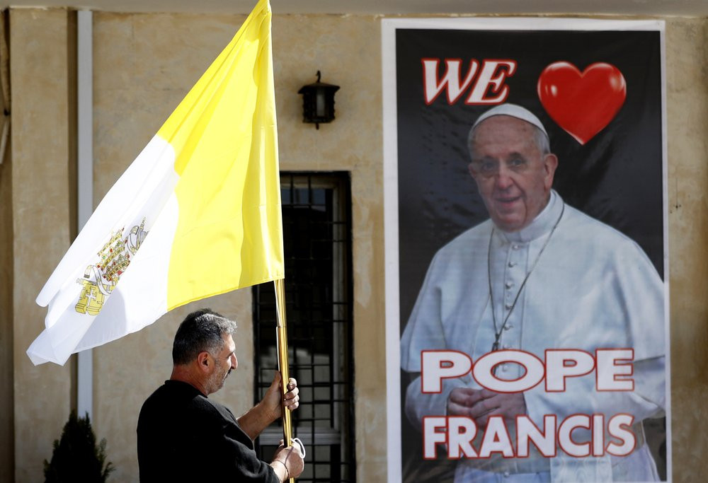 A Christian priest holds a Vatican flag as he walks by a poster of Pope Francis during preparations for the Pope's visit in Mar Youssif Church in Baghdad, Iraq, Friday, Feb. 26, 2021.