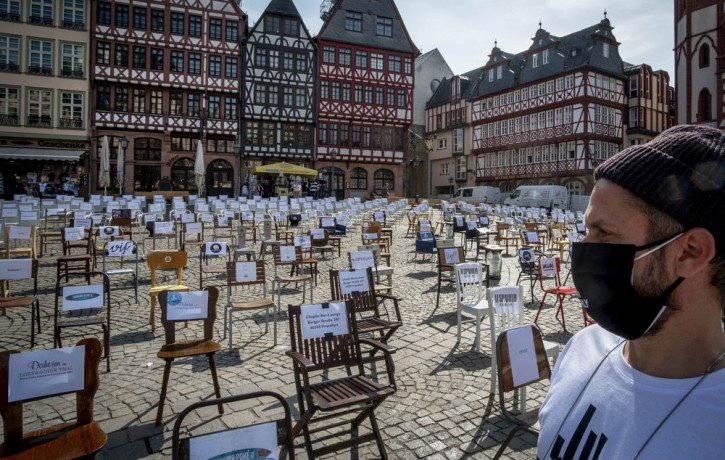 In this April 24, 2020 file photo, a man with a face mask watches empty chairs with names of bars and restaurants on the Roemerberg square in Frankfurt, Germany.